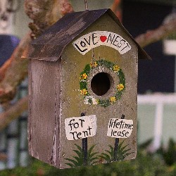 Cute homemade rustic birdhouse with love nest with a heart painted over the hole and two signs painted under the hole.  One says for rent and the other says lifetime lease.